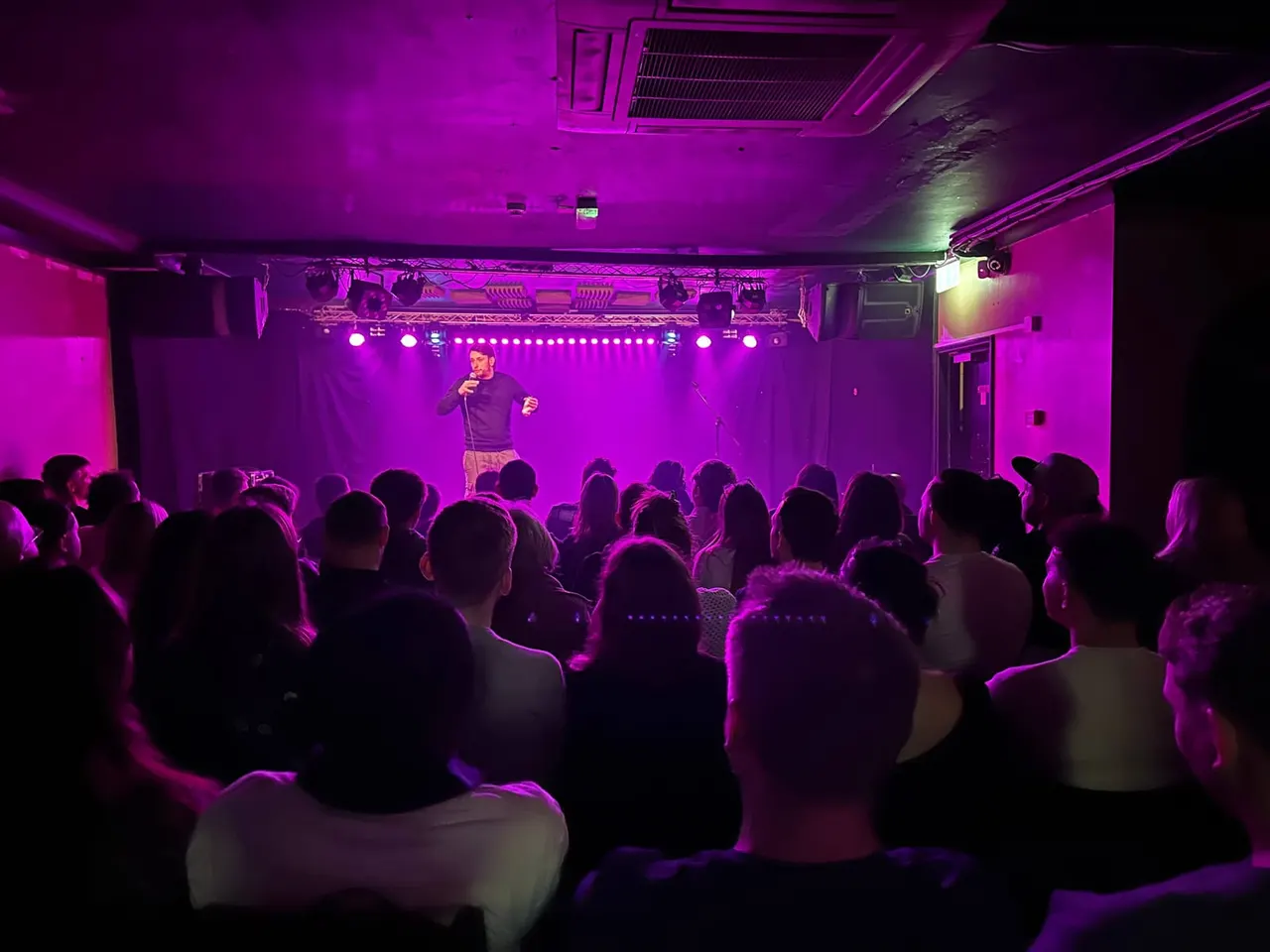 Comedian performing on stage to a seated audience in a basement venue lit with vibrant magenta lighting.