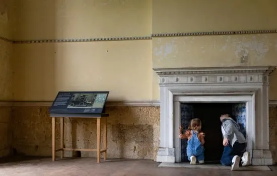 A family with young children enters a historic doorway while an informational display panel stands nearby in a bare stone chamber.