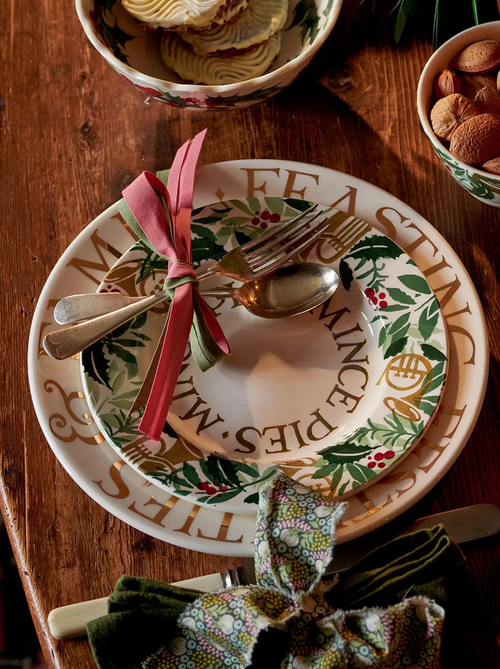 An overhead view of a festive table setting with an Emma Bridgewater plate, silverware tied with pink ribbon, bowls of biscuits and sweets on wooden surface.