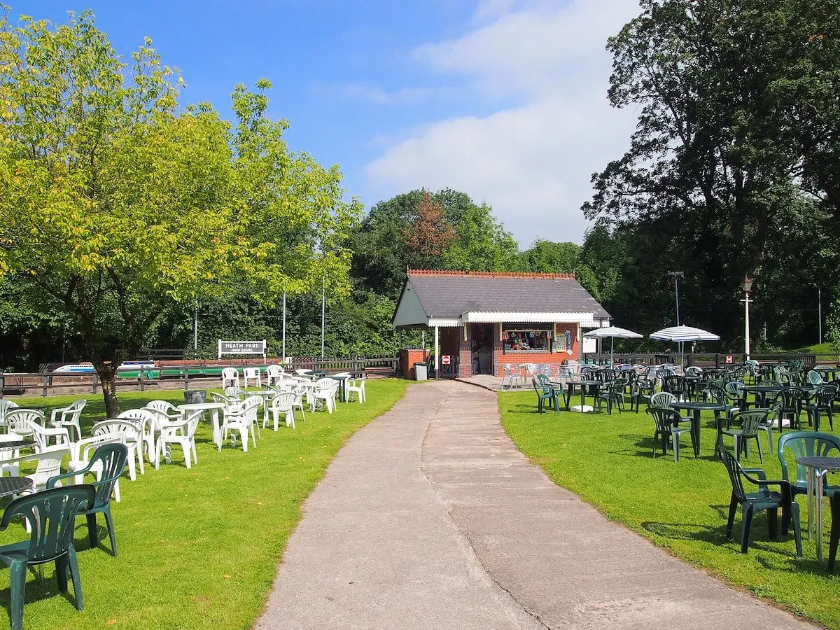 Outdoor leisure venue with pathways, seating areas, a brick building with refreshment facilities, and mature trees under blue sky.