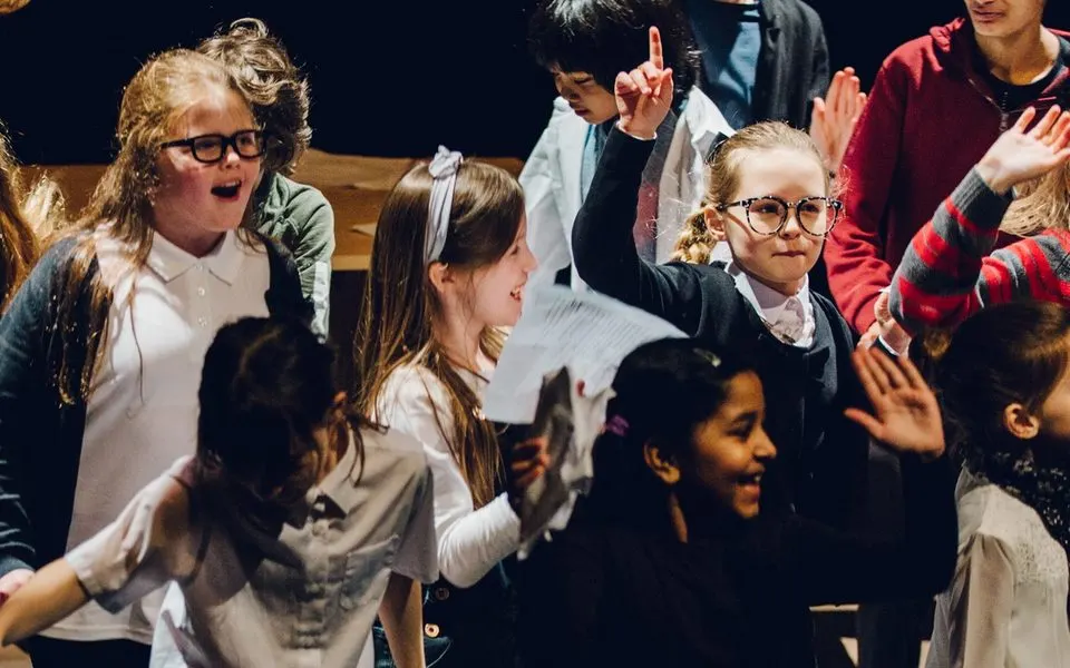 Children performing on stage, singing and dancing together with raised hands and engaged expressions.
