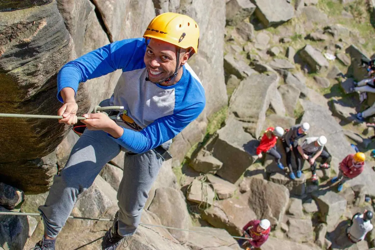A man in a yellow helmet and blue shirt rock climbing on a quarry face with spectators watching below.