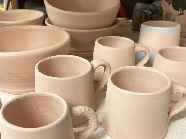 Collection of unfired cream ceramic vessels including mugs and bowls displayed on a work surface.
