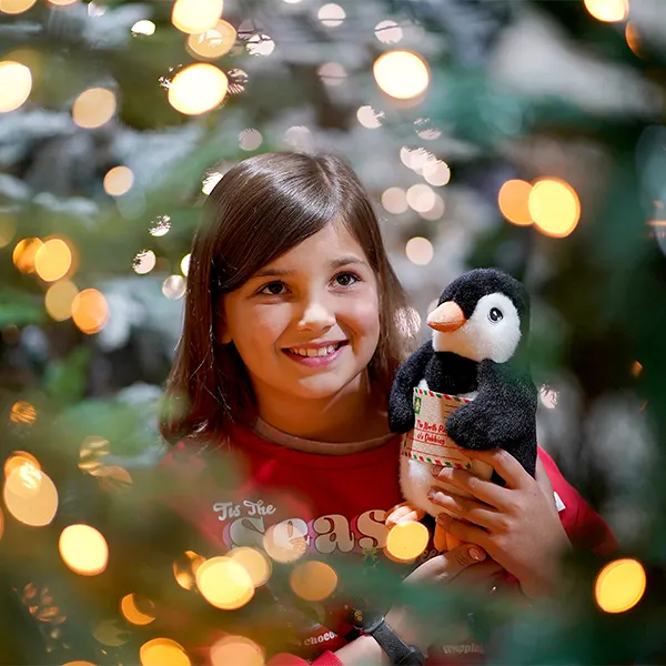 Young girl smiling at camera holding a toy penguin, surrounded by twinkling lights and evergreen foliage.