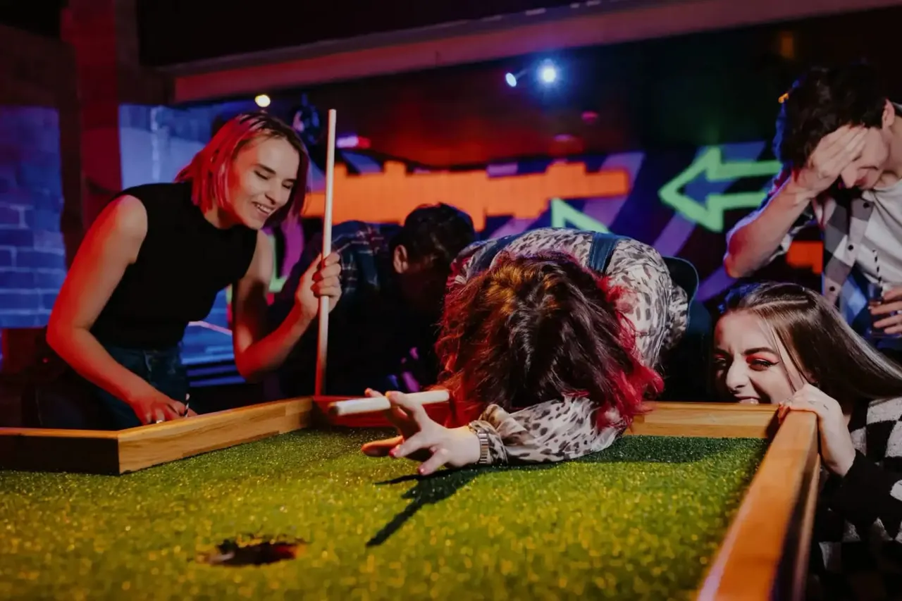 Young people playing mini golf indoors under colourful neon lighting in an entertainment venue.