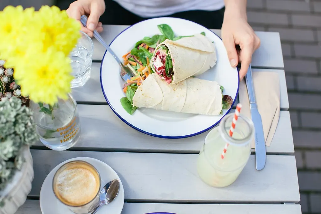 Person dining outdoors at a table with two wraps filled with salad and vegetables, alongside coffee and a drink.