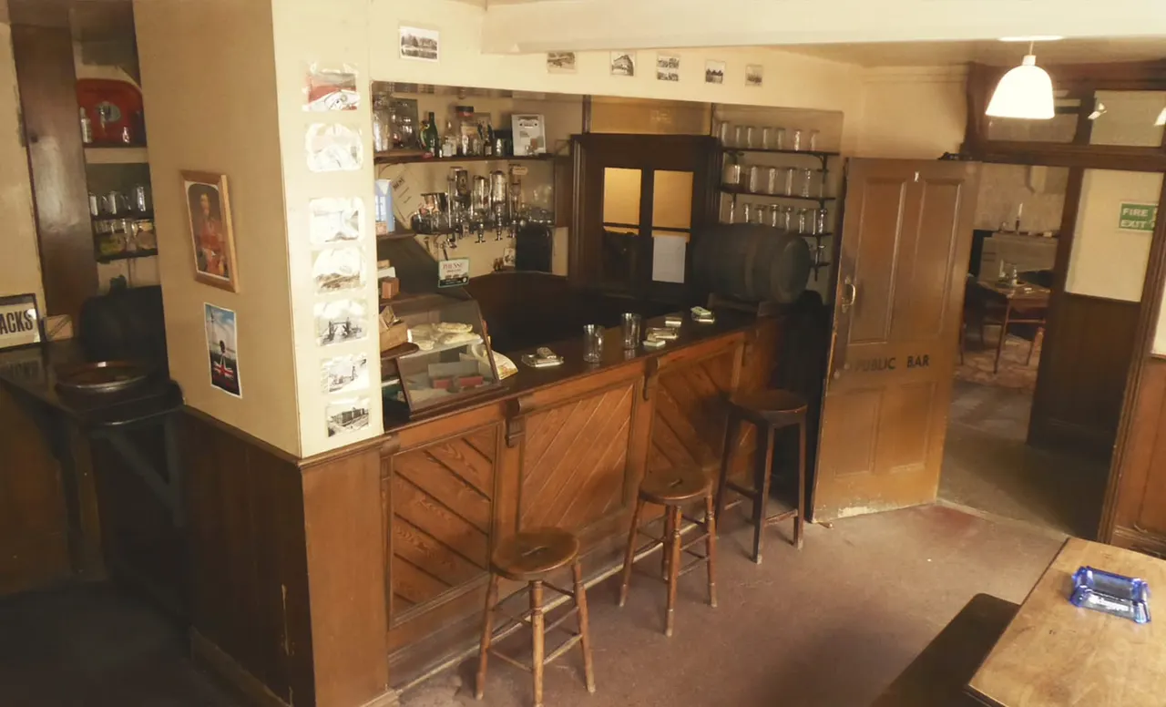 Empty pub bar interior with wooden counter, bar stools, shelves of spirits, and framed photographs on cream-coloured walls.