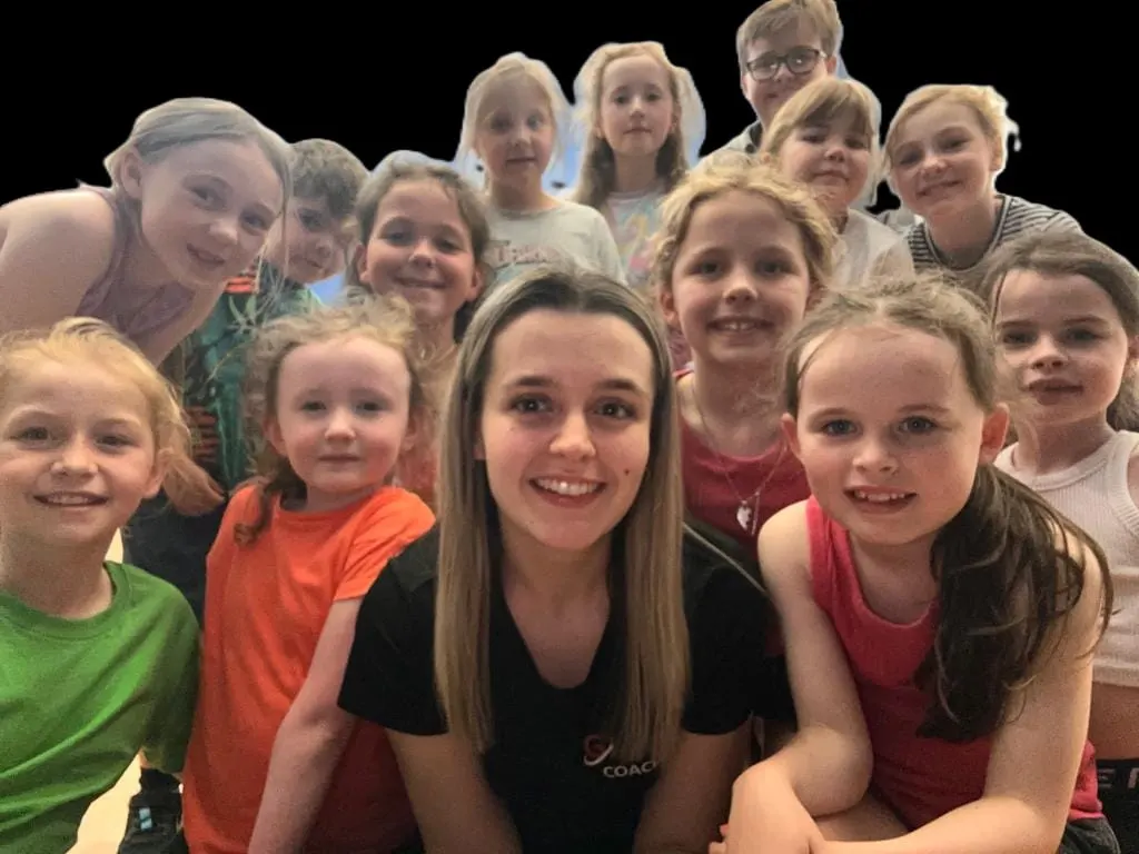A group of children and a young adult instructor smiling at the camera during a coaching session.