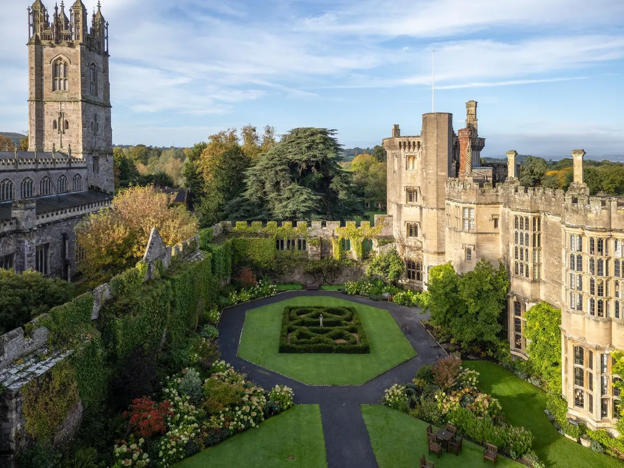 Medieval castle with formal gardens, tower, and manicured lawns surrounded by stone walls and countryside.
