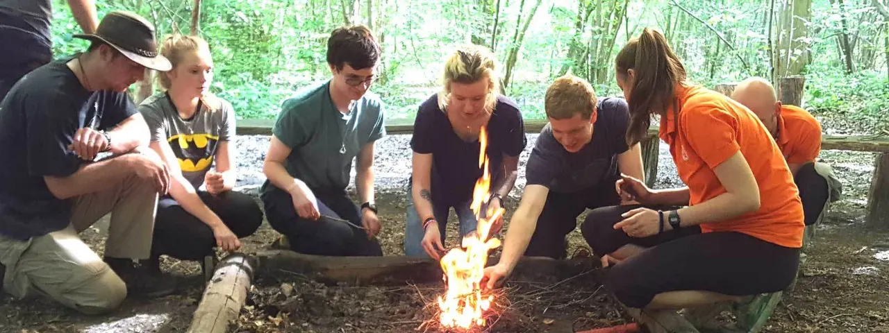 A group of adults and young people gathered around a campfire in a woodland clearing, tending to the flames together.