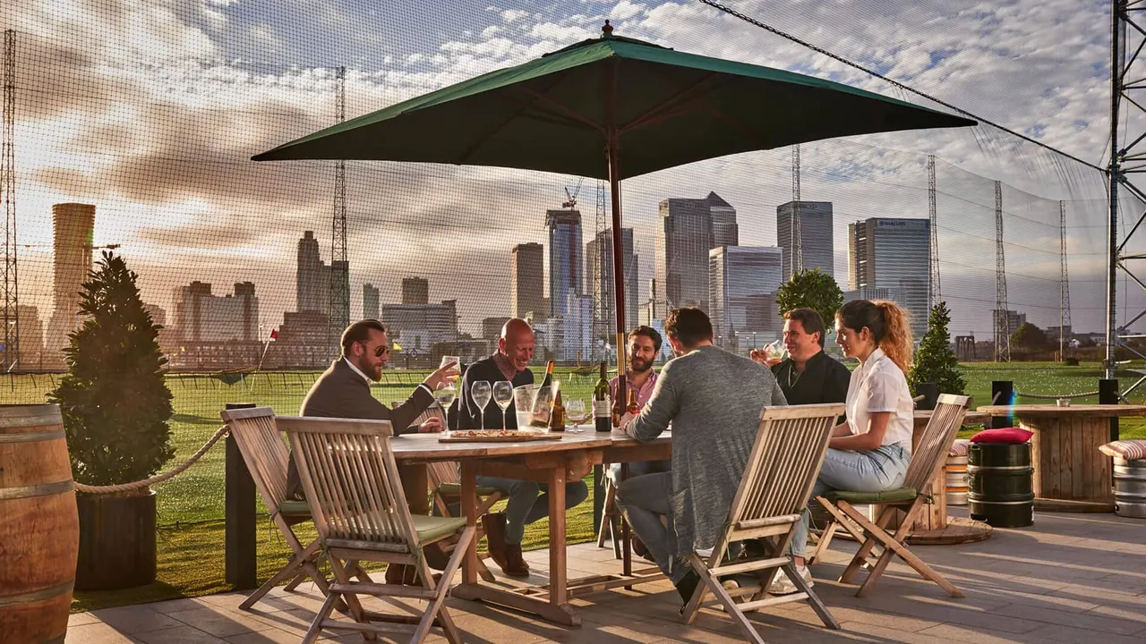 Group of six people dining outdoors under a green parasol with a city skyline visible beyond netting.