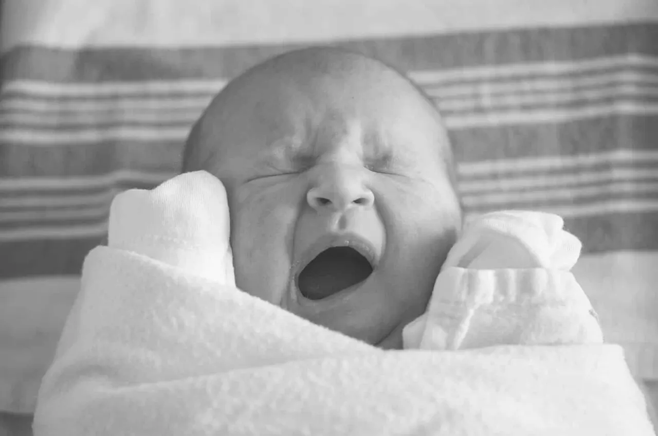 A newborn baby yawning with eyes closed, wrapped in white blankets and wearing white mittens.