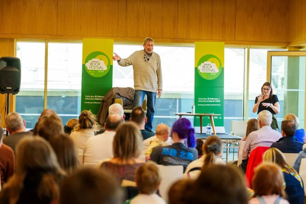 A man speaking at a book festival event to an audience seated in a modern library space with large windows.