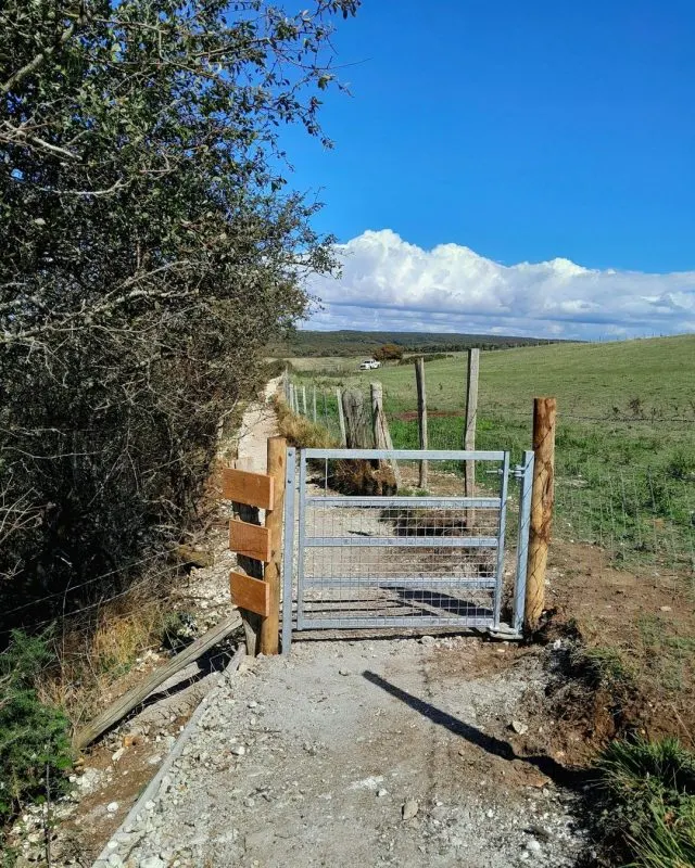 PHOTOGRAPH

Metal farm gate on a rural track with rolling green fields and hedgerow under blue sky.