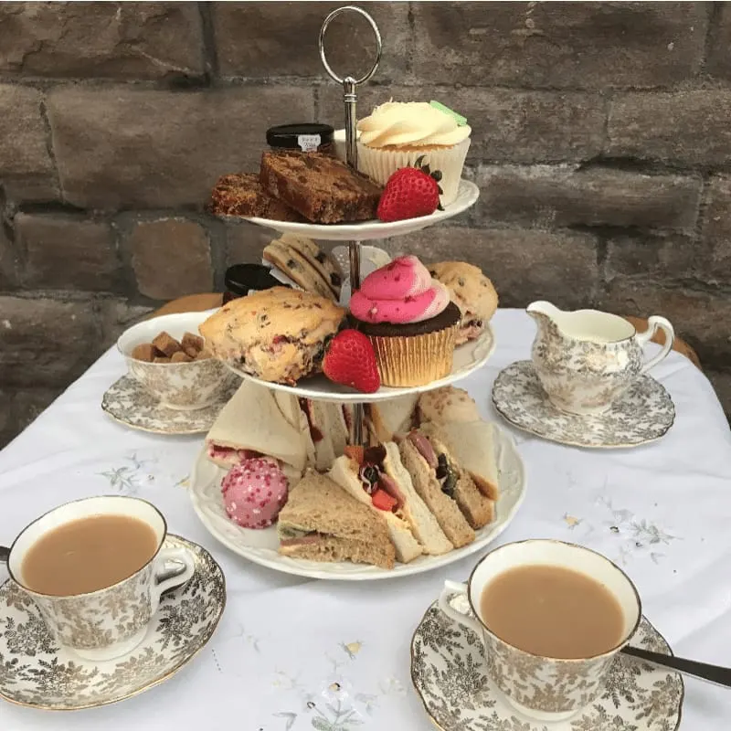 Three-tier afternoon tea stand with sandwiches, cakes, and pastries, surrounded by patterned teacups and saucers against a brick wall.