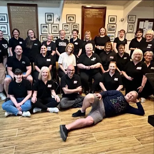 Large group of adults in matching black shirts posing together indoors, with one person lying on the floor in the foreground.