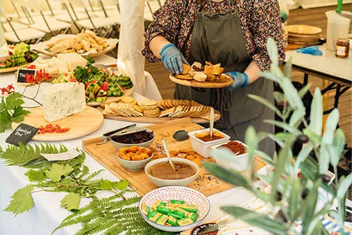Person in apron and gloves preparing food at a buffet table with salads, breads and various dishes.