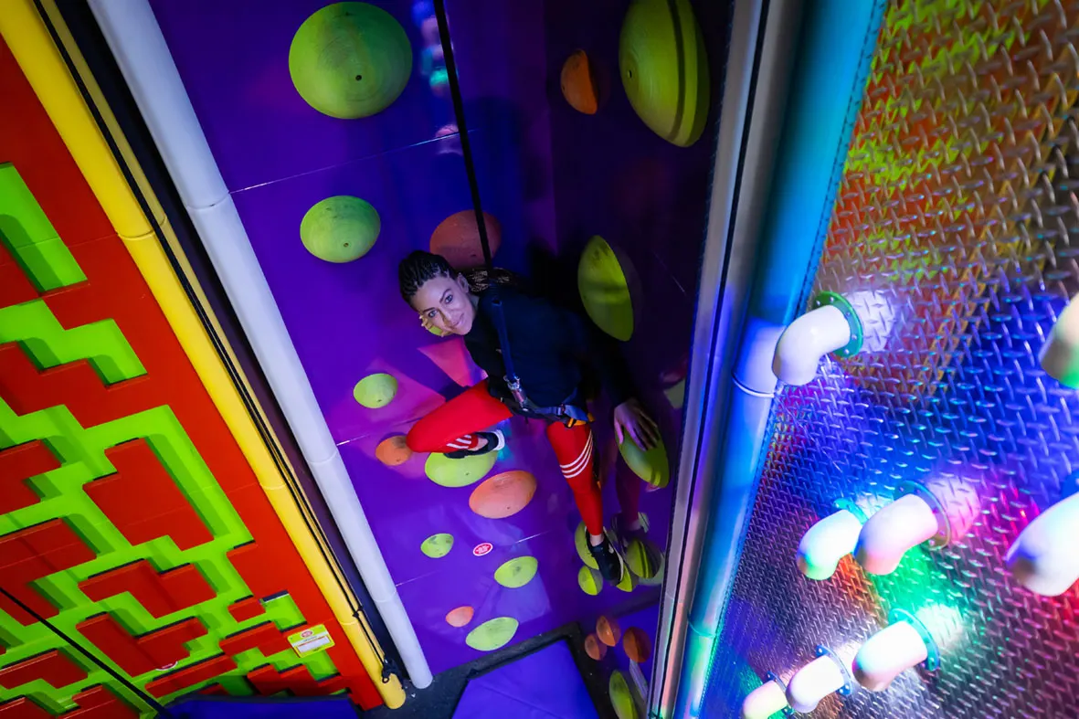 Child climbing an indoor wall with illuminated hand and foot holds in a brightly lit climbing centre.