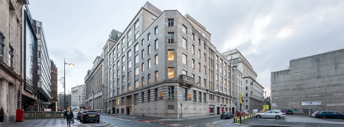 Grand Victorian stone building with lit windows at dusk, surrounded by modern city streets and parked cars.