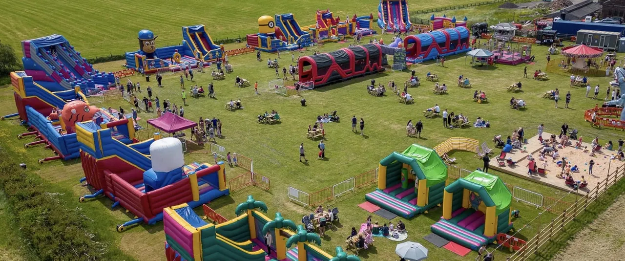 Aerial view of a large outdoor inflatable play park with colourful bouncy castles and slides on a grass field, with families and children enjoying activities throughout.