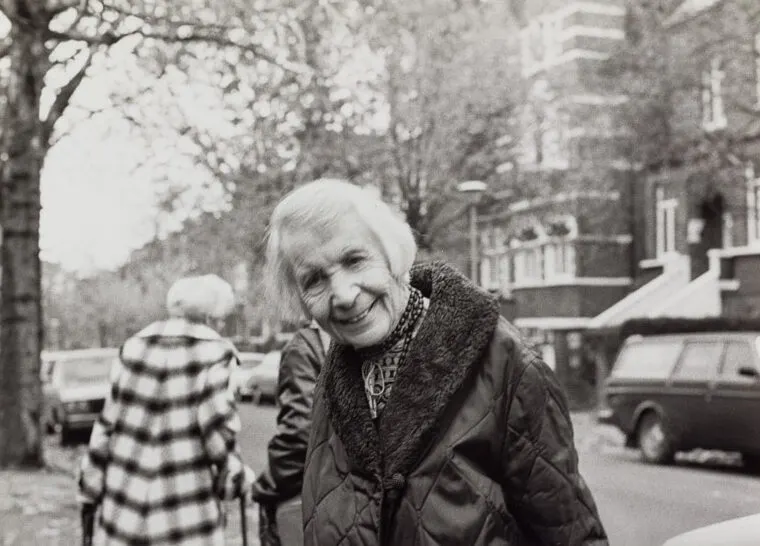 Black and white photograph of an elderly woman smiling at the camera on a tree-lined residential street, with another person visible in the background.