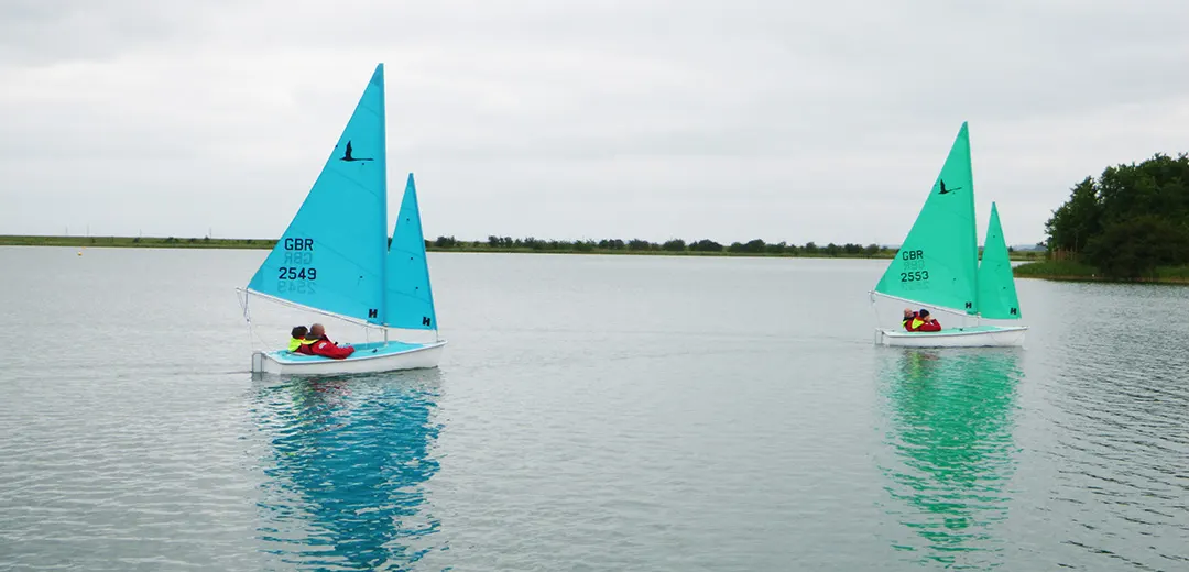 Two small sailing boats with bright turquoise and green sails on calm water, each with two people aboard.
