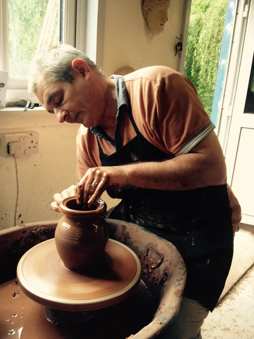 Older man working clay on a potter's wheel, shaping a terracotta vessel in a bright studio.