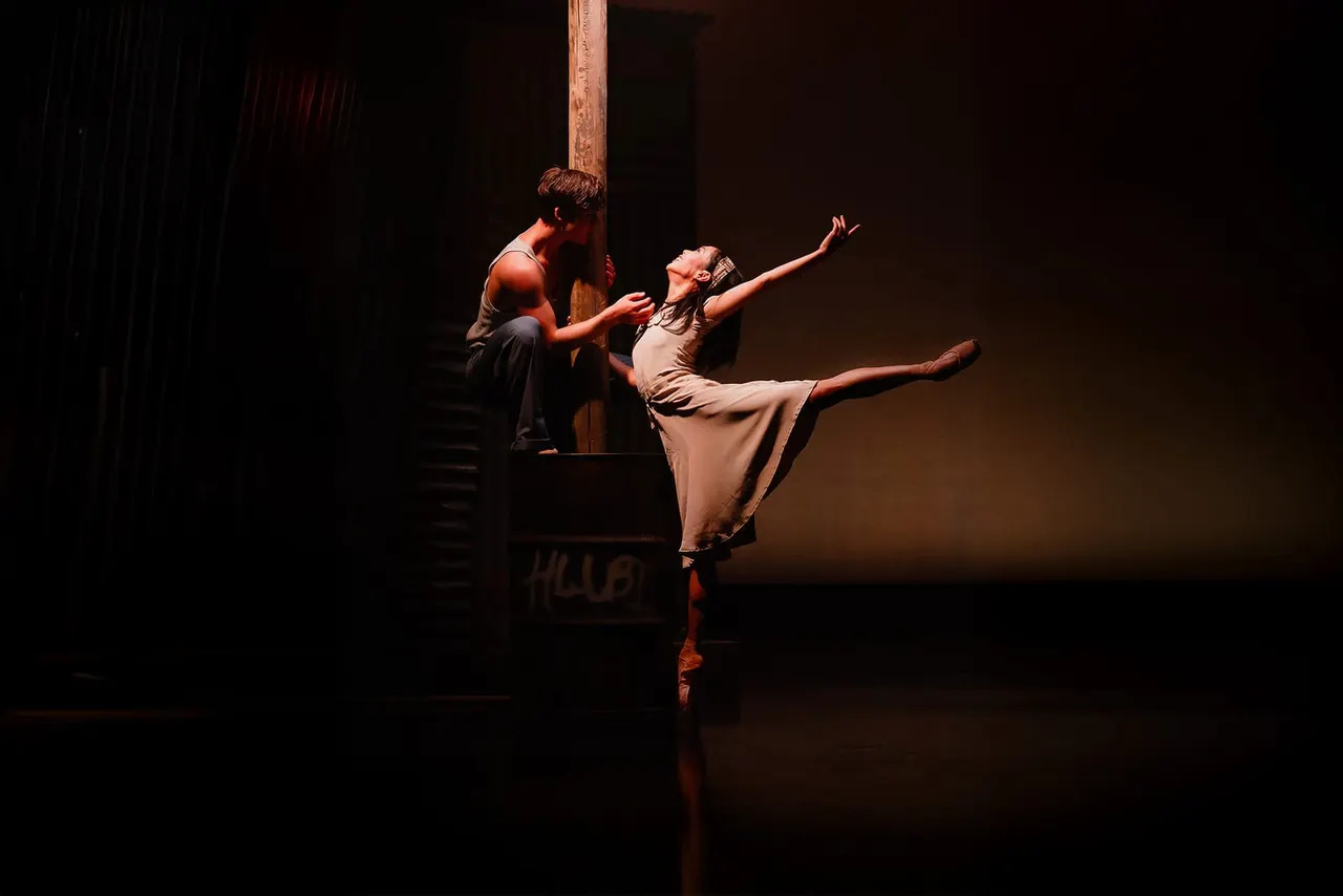 Two ballet dancers perform on stage, one in pointe shoes executing a leg extension whilst the other provides support.