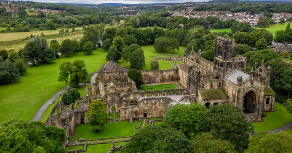 Aerial view of ruined medieval abbey surrounded by green lawns and trees, with residential areas visible in the distance.