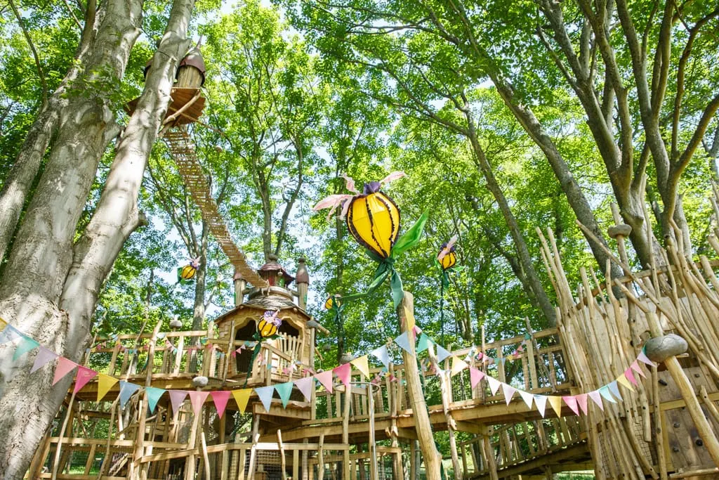 Wooden treehouse structure with platforms and walkways decorated with bunting and large colourful flower sculptures, nestled among tall trees.
