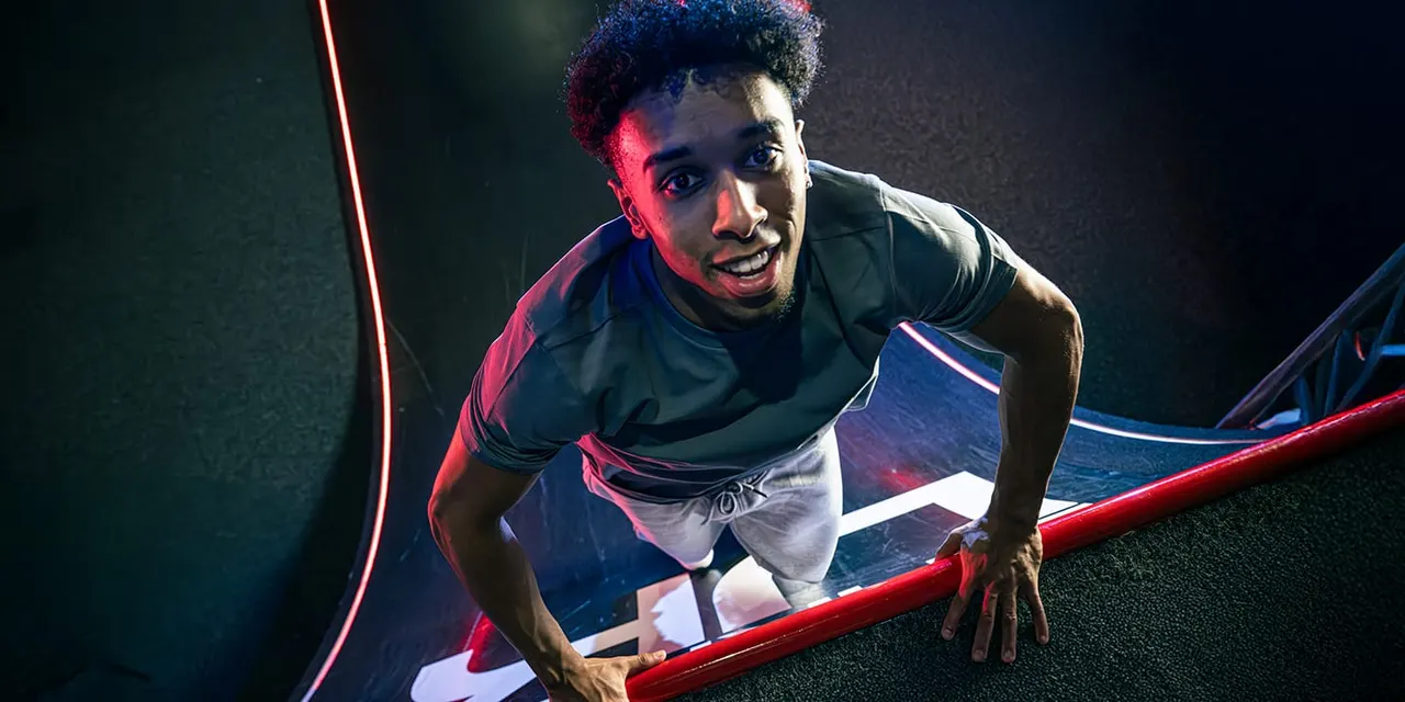 Person climbing over a red obstacle bar during an indoor ninja warrior course, lit with neon lighting.