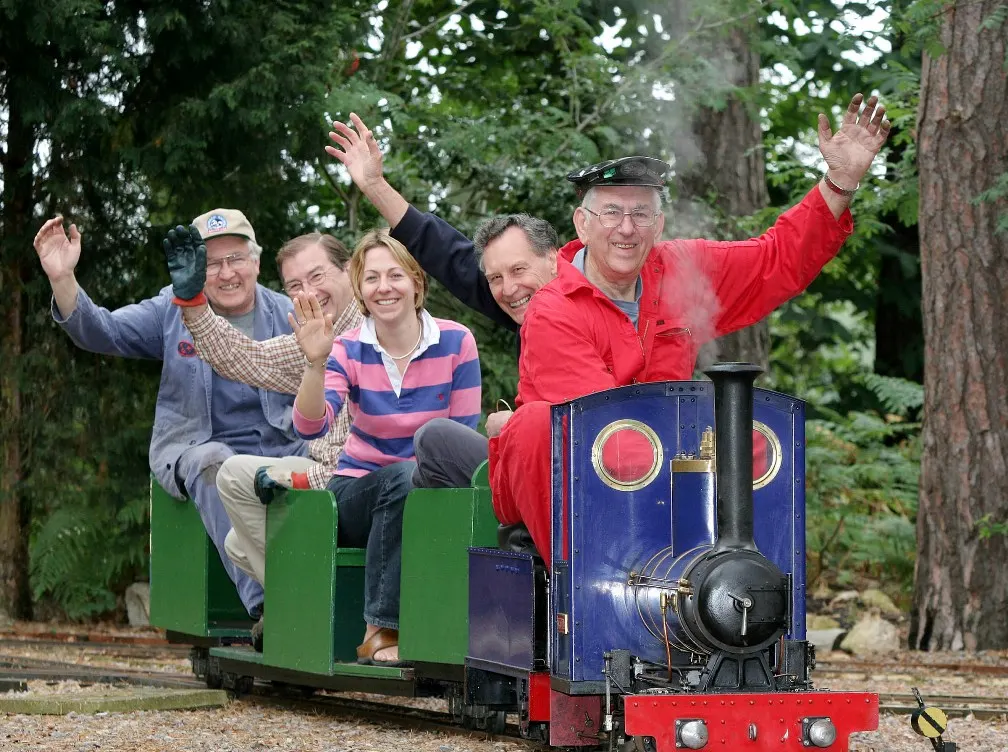 Five adults waving and smiling whilst riding in a carriage towed by a miniature steam locomotive through tree-lined tracks.