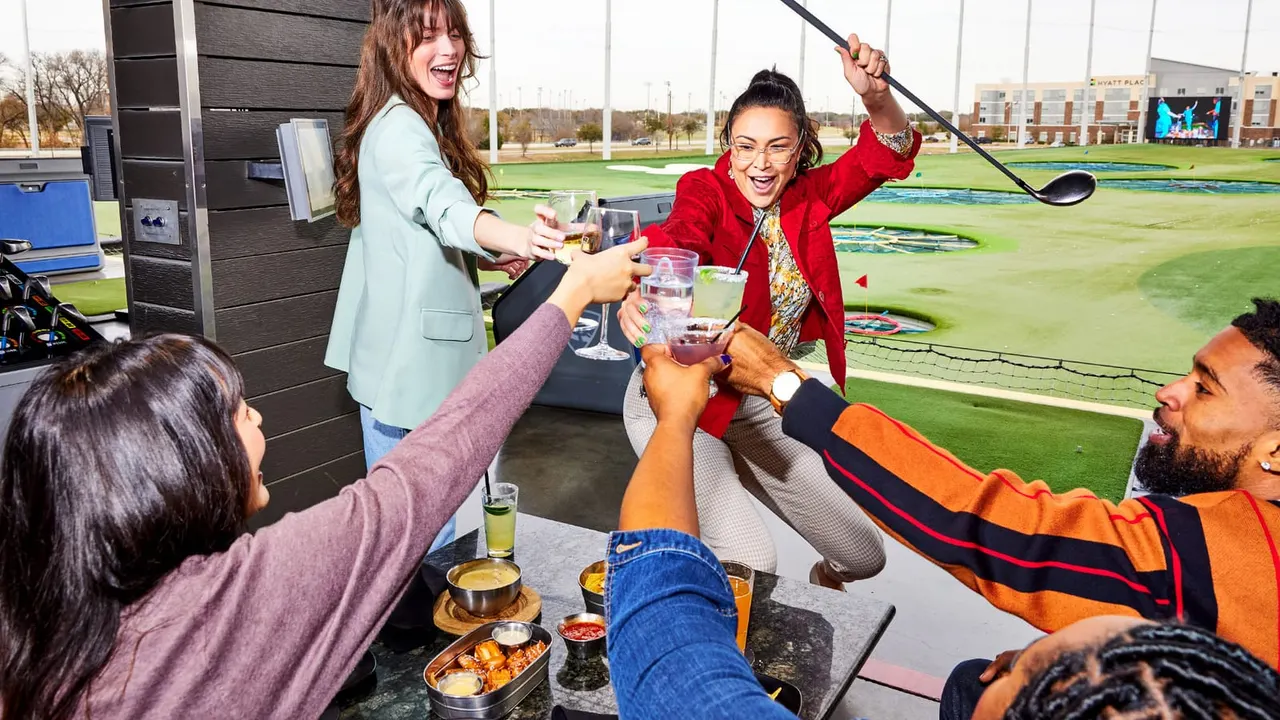 Group of people toasting with drinks at an indoor golf bay overlooking a driving range.