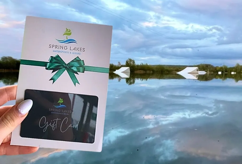 Hand holding a Spring Lakes gift card with green bow, lakeside setting with calm water and moored boats behind.