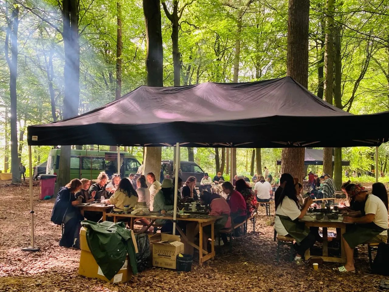 Groups of visitors seated at wooden tables under a black canopy in a woodland clearing surrounded by tall trees.