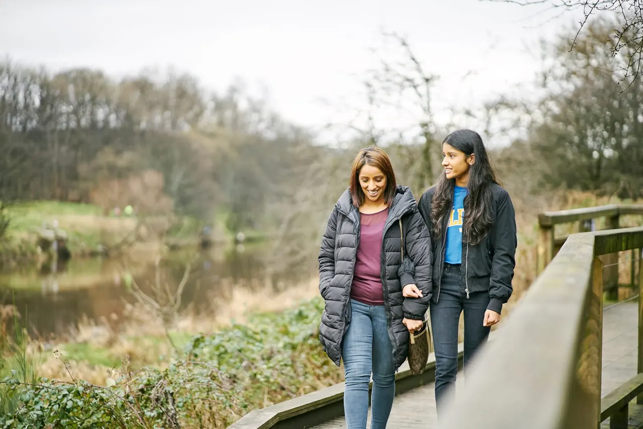 Two young women walk along a wooden boardwalk path overlooking a river valley with bare winter trees.