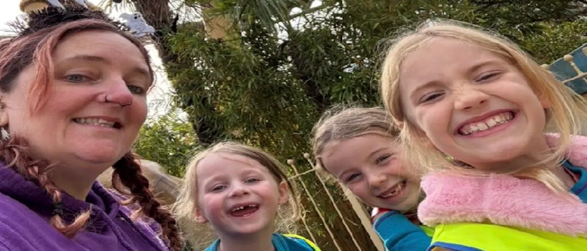 An adult and three children smiling at the camera outdoors amongst trees and greenery.