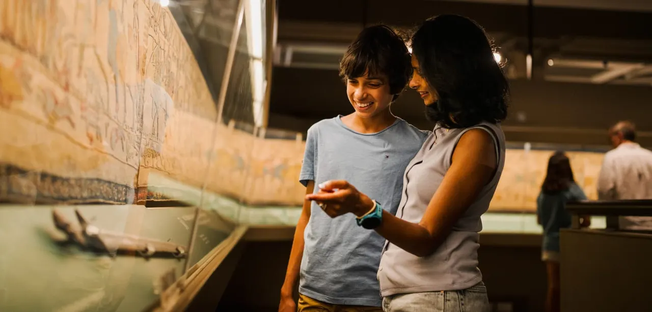 Two young people smiling whilst viewing a large stone monument indoors at an archaeological site.