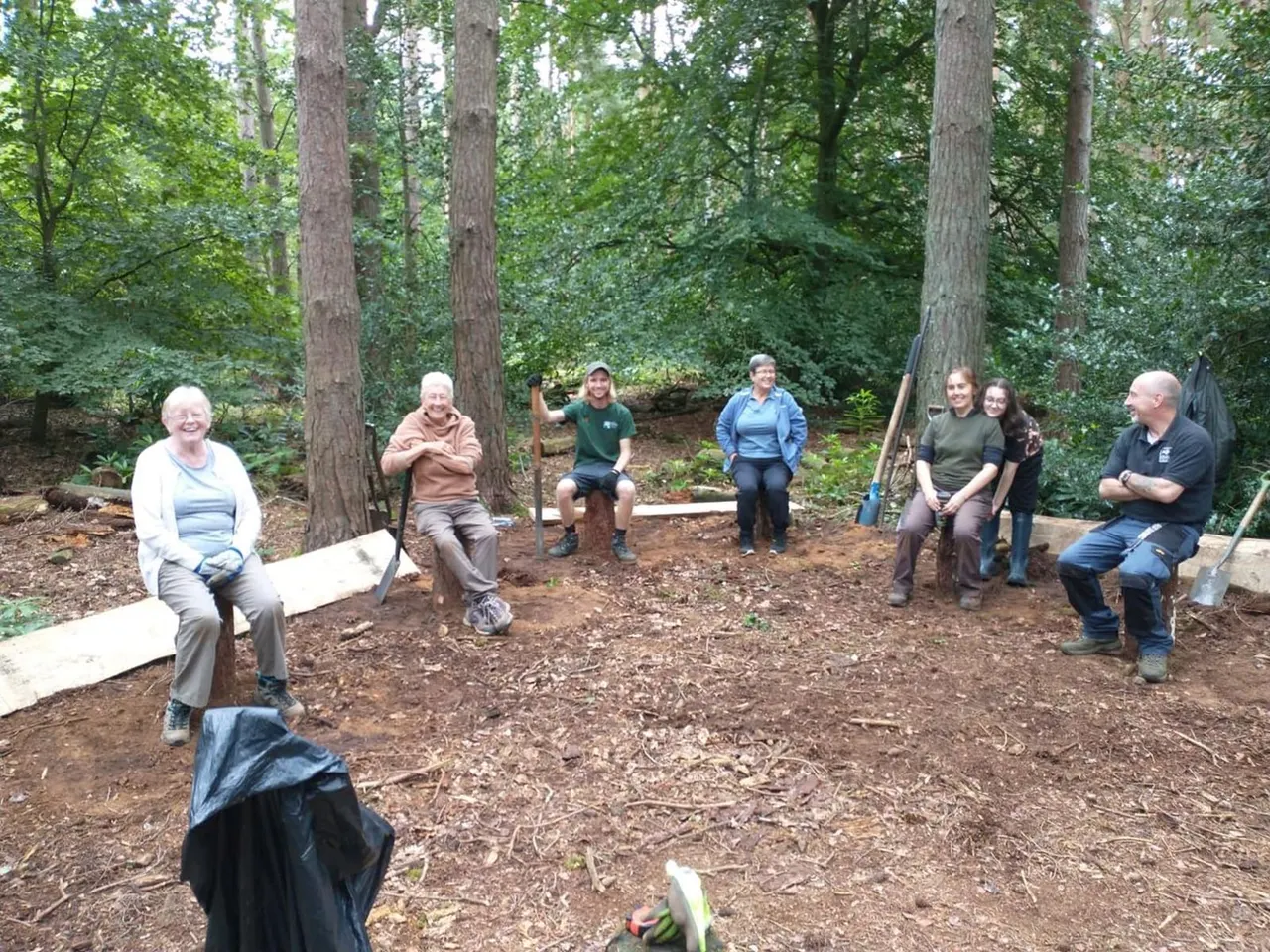 Group of visitors sitting on wooden benches in a woodland clearing surrounded by tall trees.
