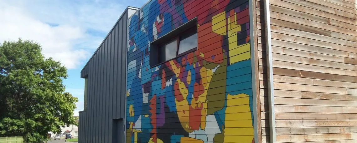 A modern building with colourful abstract mural artwork on its blue-painted corrugated metal wall, next to wooden cladding.