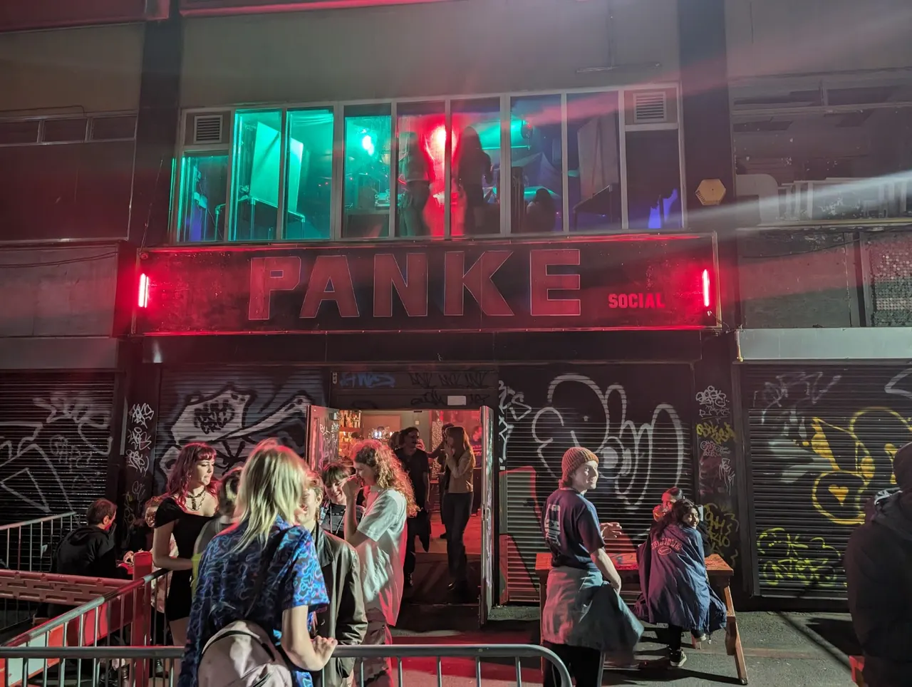 People gather outside a nightclub venue with neon red and cyan lighting at its entrance and windows.