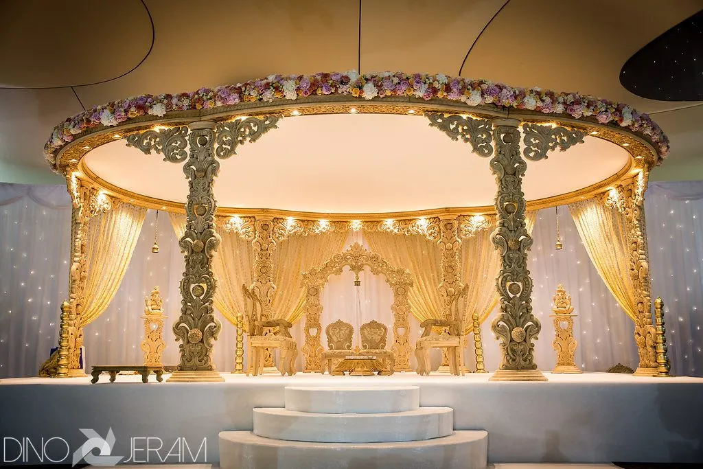 Ornate golden mandap with floral garland, elaborate carved columns, and illuminated drapery for a wedding ceremony.