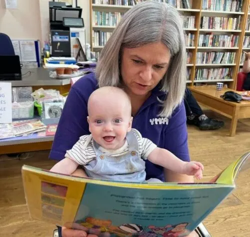 An adult and baby reading a picture book together in a library surrounded by bookshelves.