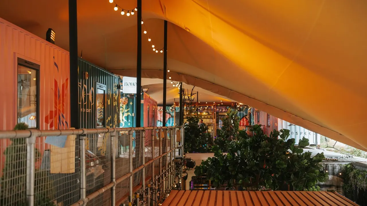 Covered market or leisure venue with colourful shipping container retail units, string lights, potted plants, and metal railings along a wooden deck at dusk.