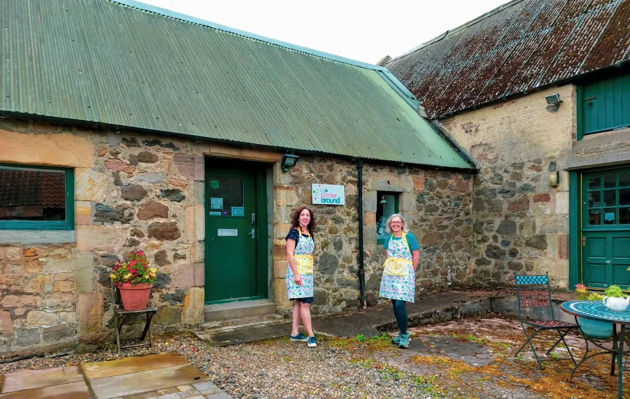 Two women in matching aprons stand in a courtyard of stone farm buildings with green doors and corrugated metal roofing.