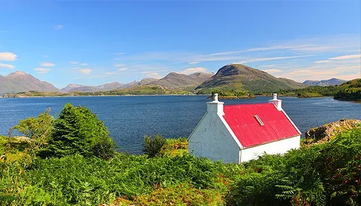 A white cottage with a bright red roof sits beside a blue loch with mountains beyond.
