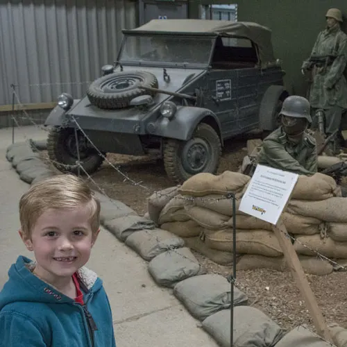 Young child smiling at camera beside military museum display with vintage army vehicle and uniformed figures.