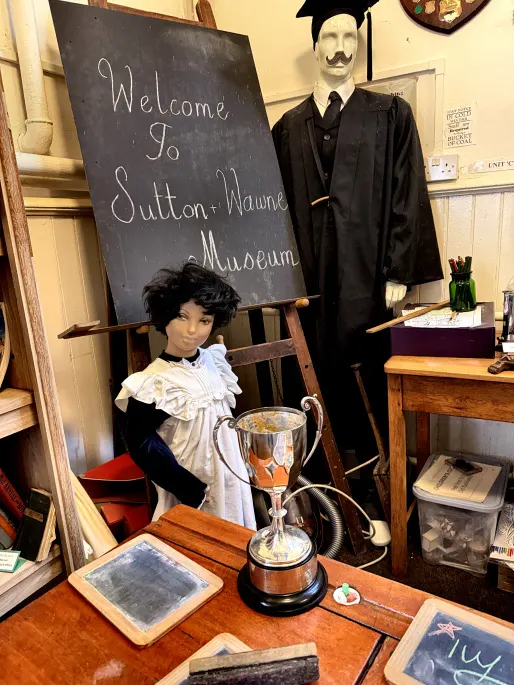 Museum display featuring mannequins dressed in period costume with a welcome sign, trophy, and historical artefacts on wooden furniture.