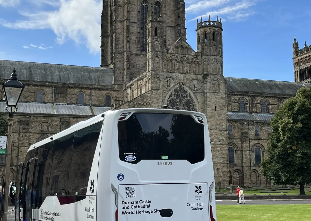 Accessible shuttle bus parked in front of Durham Cathedral, a large stone Gothic building with towers and ornate windows.