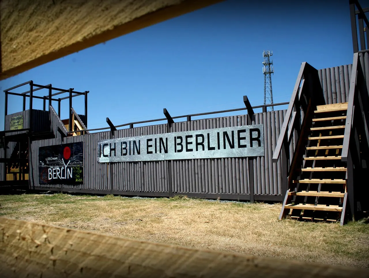 Corrugated metal structures with "Ich Bin Ein Berliner" signage and wooden stairways at an outdoor paintball venue under clear blue sky.